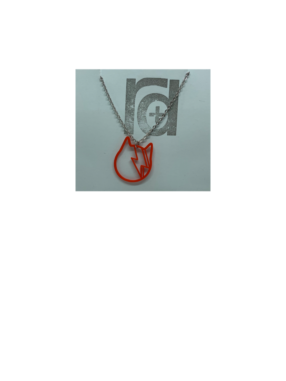 On a light blue R+D necklace card is a stainless steel chain with a red 3D printed pendant. The pendant is printed using a plant based filament. It is the shape of a cat with a lightning bolt across its face to mimic David Bowie's iconic album cover. 